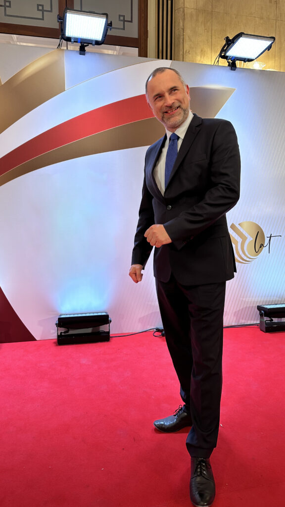 Paweł Kurpiewski stoi na czerwonym dywanie w garniturze, z tylu świecą reflektory a on się uśmiecha z zadowoleniem