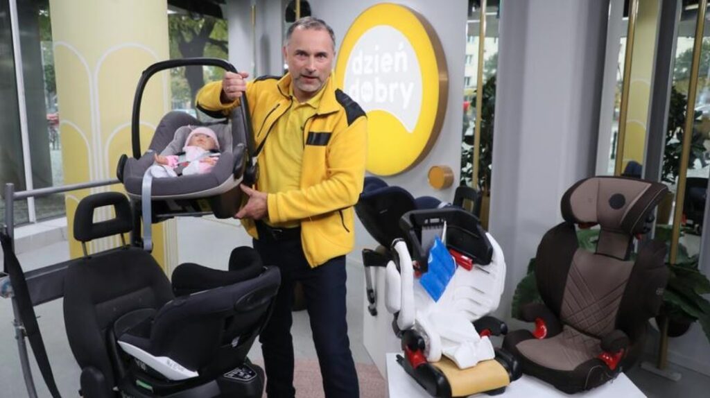 Kurpiewski w TVN mowi o fotelikach samochodowych, trzymajac jeden w ręku. Ubrany jest w żółtą koszulkę i polar